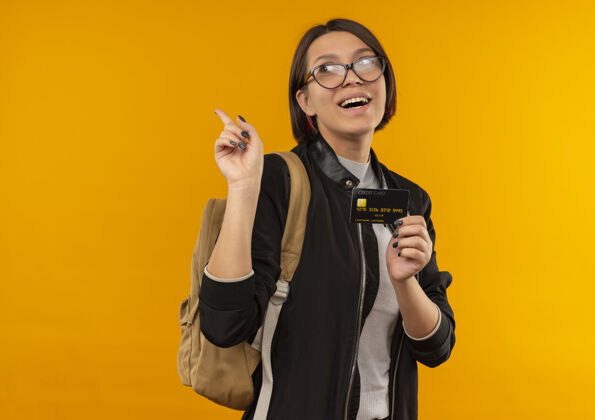 手指快乐的年轻学生女孩戴着眼镜 背着信用卡看着手袋边举起手指孤立在橙色包壁板眼镜