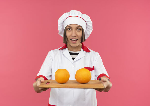 橙色身着厨师制服的快乐年轻女厨师伸出插有橘子的砧板 上面是粉红色的板子伸展粉色