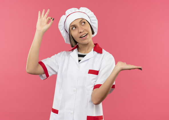 粉红快乐的年轻女厨师穿着厨师制服做着“ok”的手势 空手看着粉红色的隔离边表演空烹饪