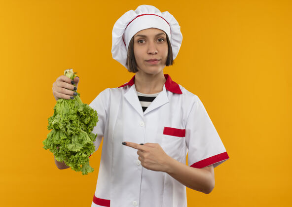 年轻身着厨师制服的年轻女厨师手拿莴苣 指着橙色背景上孤立的镜头厨师拿着生菜