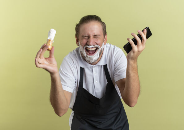 喜悦快乐的年轻英俊的理发师穿着制服拿着手机和剃须刷闭着眼睛 用剃须膏涂抹在橄榄绿的胡子上年轻橄榄油刷子