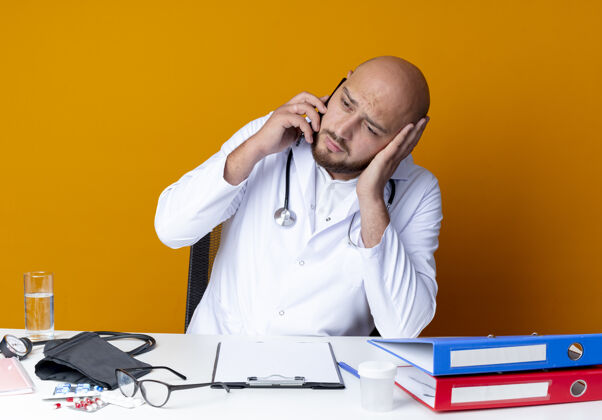 秃头悲伤的年轻秃头男医生穿着医用长袍和听诊器坐在办公桌旁 拿着医疗工具 在橙色背景下孤立地讲着电话坐着说话听诊器