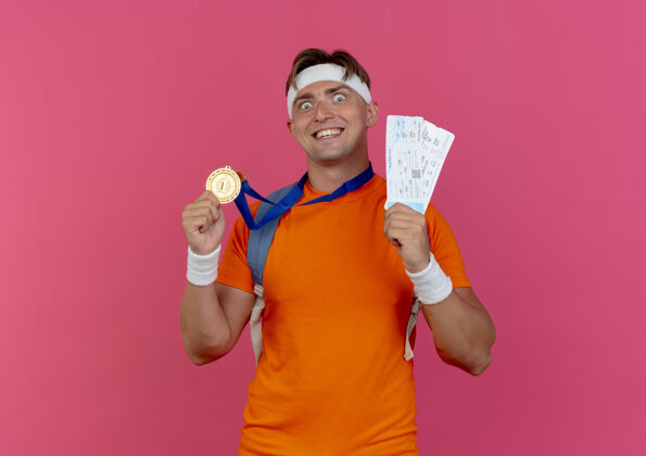 欢乐快乐的年轻帅气的运动型男人戴着头带和腕带 背上包着奖章 脖子上拿着飞机票 奖章被隔离在粉色上票年轻背部