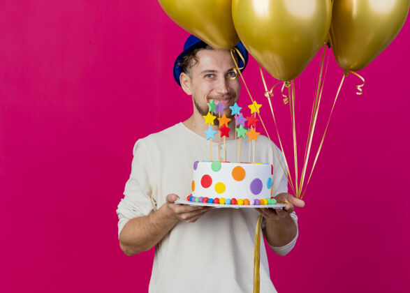 人高兴的年轻英俊的斯拉夫党家伙戴着党的帽子拿着气球和生日蛋糕与明星看前面的粉红色墙上孤立的复制空间姿势聚会帅气