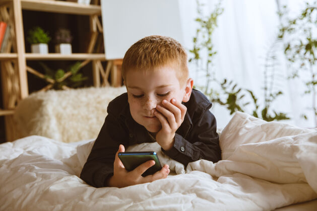 肖像男孩在家里使用不同的小玩意带智能手表 智能手机或平板电脑和耳机的小模型自拍 聊天 玩游戏 看视频孩子和现代科技的互动无线移动小学生
