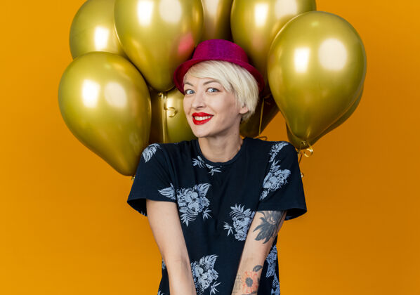 帽子微笑着的年轻金发派对女郎戴着派对帽站在气球前看着橙色墙上孤立的前方人聚会金发