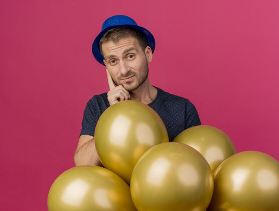脸失望的帅哥戴着蓝色的派对帽 手里拿着氦气球 隔离在粉红色的墙上 还有复制空间帅哥穿聚会