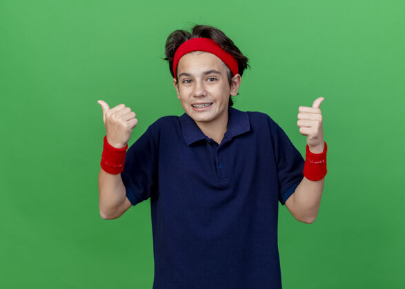人面带微笑的年轻帅气的运动男孩戴着头带和戴着牙套的腕带看着前面 竖起大拇指孤立地站在绿色的墙上 留着复制空间腕带头带姿势