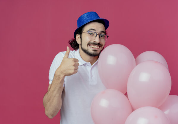气球笑容可掬的帅哥戴着眼镜 戴着蓝色的帽子 手里拿着气球 大拇指孤立地竖起 背景是粉色的微笑拿着眼镜