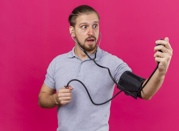 血压计年轻英俊的斯拉夫病人戴着听诊器 用血压计测量他的血压 看着隔离在粉红色墙上的复制空间男人感情立场