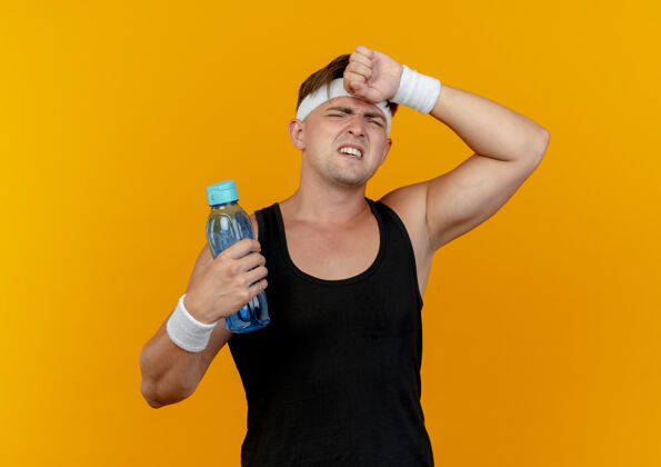 手疲惫的年轻帅气的运动型男人戴着头带和腕带拿着水瓶 把手放在孤立在橙色背景上的额头上头带瓶子抱着