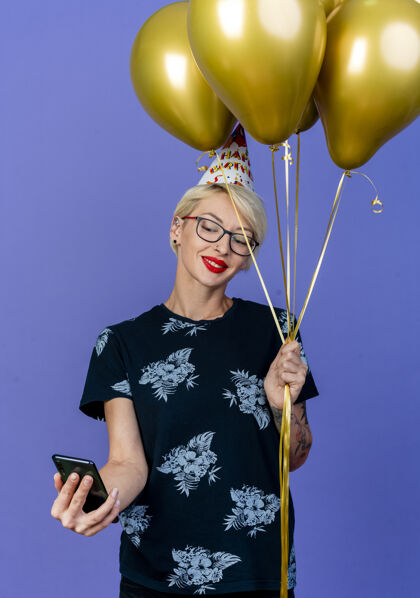 人微笑的金发女郎戴着眼镜 戴着生日帽 拿着气球和手机 独自在紫色的墙上自拍人自拍聚会