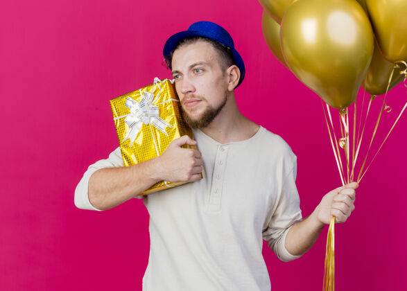 礼物年轻帅气的斯拉夫派对男戴着派对帽拿着气球 拿着礼盒摸着脸看着粉色墙上孤立的一面衣服气球帅气