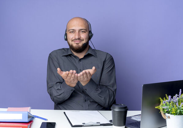 工具高兴的年轻秃头呼叫中心男子戴着耳机坐在办公桌上 工作工具显示空手在相机隔离紫色背景手中心秃头