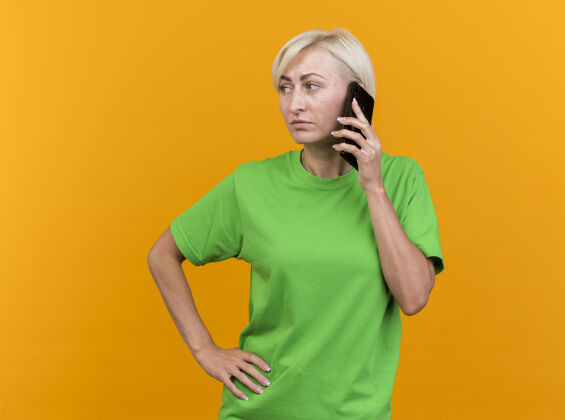 斯拉夫自信的中年金发斯拉夫女人在打电话 手放在腰上 看着隔离在黄色墙壁上的一面 还有复制空间表情衣服壁板