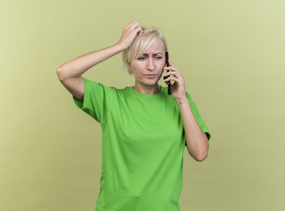 立场皱眉的中年金发斯拉夫女人在打电话 看着一边把手放在头上 隔离在橄榄绿的墙上 留着复印空间感情手人