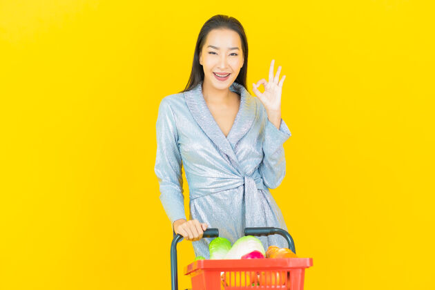 杂货店一幅美丽的亚洲年轻女子微笑的画像 黄色的墙上挂着超市的购物篮零售市场女性