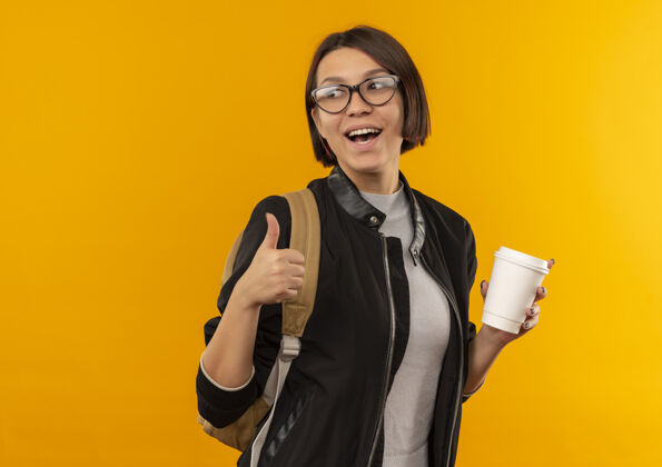 杯子快乐的年轻学生女孩戴着眼镜 背着袋子 拿着塑料咖啡杯 一边看一边竖起大拇指 孤立地站在橘子上年轻咖啡表演