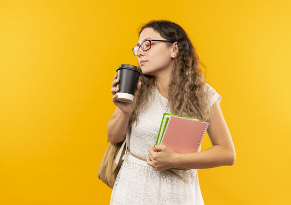垫年轻漂亮的女学生戴着眼镜 背着书包 拿着笔记本 嗅着咖啡杯 闭着眼睛 隔离在黄色背景上 留着复印空间穿背部女学生