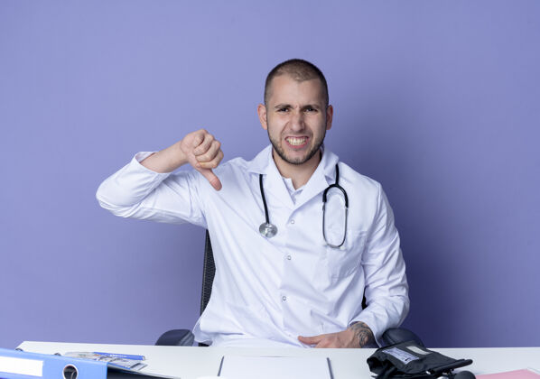 坐着穿着医用长袍和听诊器的年轻男性医生坐在办公桌旁 工作工具显示拇指朝下 孤立在紫色背景上工具医生表演