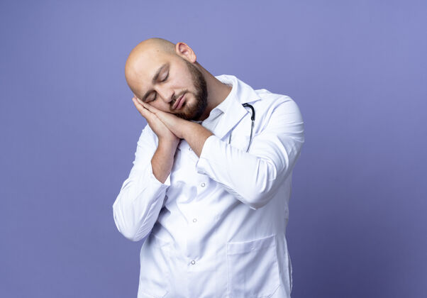 男性年轻的秃头男医生闭着眼睛 穿着医用长袍和听诊器 在蓝色背景上显示睡眠姿势手势年轻闭嘴