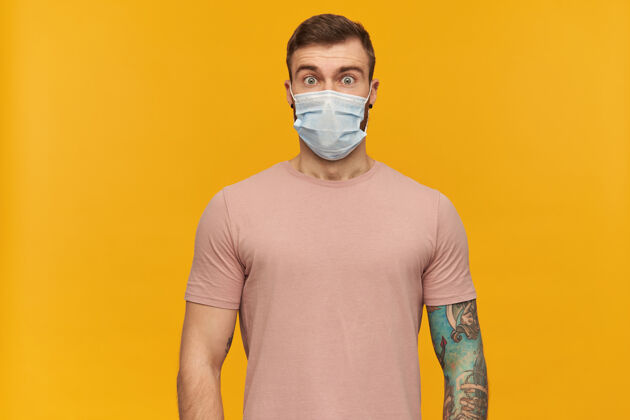 健康惊讶的纹身年轻人穿着粉色T恤 戴着卫生口罩 防止胡须感染 惊讶的看着前面的黄色墙壁肖像流感相机
