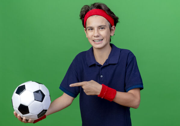 帅哥快乐的年轻帅气的运动男孩戴着头带和带牙套的腕带看着前面拿着足球 指着隔离在绿色墙壁上的足球衣服足球头带
