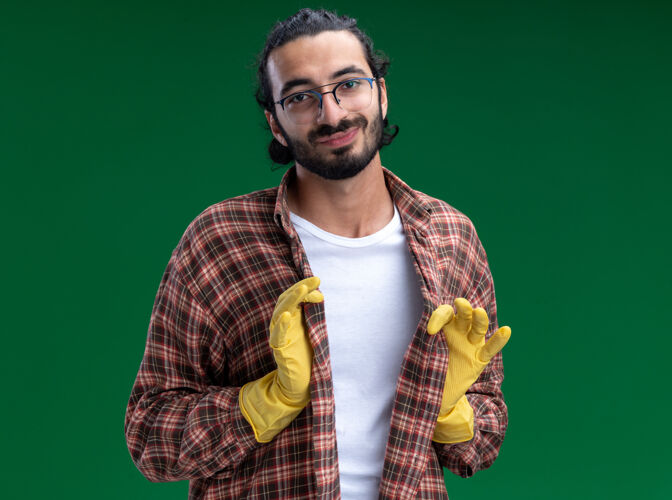 手套自信的年轻帅气的清洁工穿着t恤 戴着手套 把t恤隔离在绿色的墙上抱着小伙子穿上