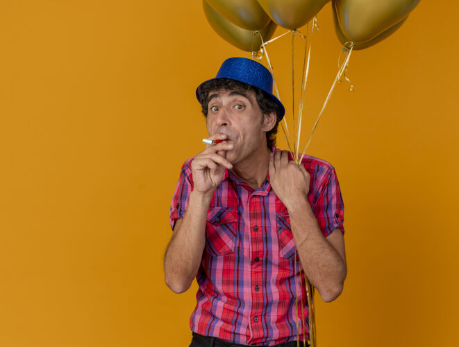 印象印象深刻的中年白种人党男子戴着党帽手持气球看着相机手持党吹风机在嘴里孤立的橙色背景与复制空间复制中年男人