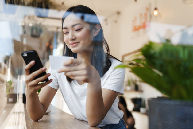 城市时尚的亚洲女孩坐在靠近窗户的咖啡馆里 用信用卡支付网上购物的费用支付卡布奇诺时尚