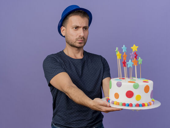 男人笨拙英俊的白人男子戴着蓝色的帽子拿着生日蛋糕看着紫色背景上的复制空间隔离帽子不知所措紫色