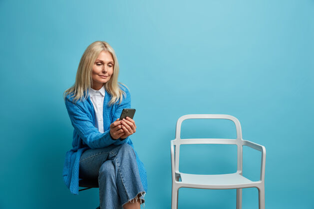 空迷人的中年妇女坐在橱柜旁排队 靠着蓝色的墙壁在椅子上摆姿势 用智能手机成人房子养老金