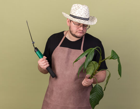 悲伤悲伤的年轻男园丁戴着园艺帽 拿着剪子 看着隔离在橄榄绿墙上的植物剪子花园帽子