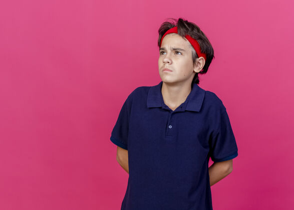 空间体贴的年轻帅气的运动男孩戴着头带和护腕 戴着牙套 双手放在背后 看着在深红色背景上与复制空间隔离的一侧年轻运动男孩