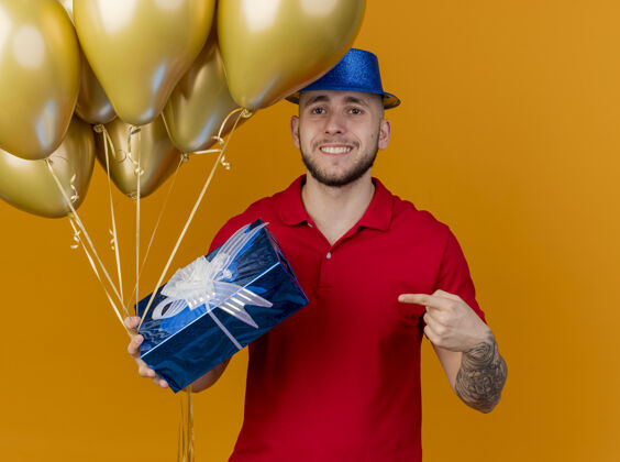 气球面带微笑的年轻帅哥斯拉夫党人戴着党的帽子拿着气球和礼品包看着相机指着礼品包孤立的橙色背景小伙子礼物派对