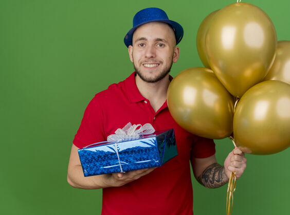 包面带微笑的年轻帅哥斯拉夫党的家伙戴着党的帽子拿着气球和礼品包看着相机隔离在绿色背景与复制空间礼物家伙绿色