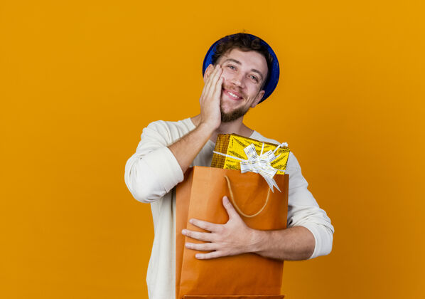 脸快乐的年轻帅哥斯拉夫党家伙戴着党的帽子拿着礼品盒在纸袋保持手在脸上橙色背景与复制空间隔离家伙手礼物