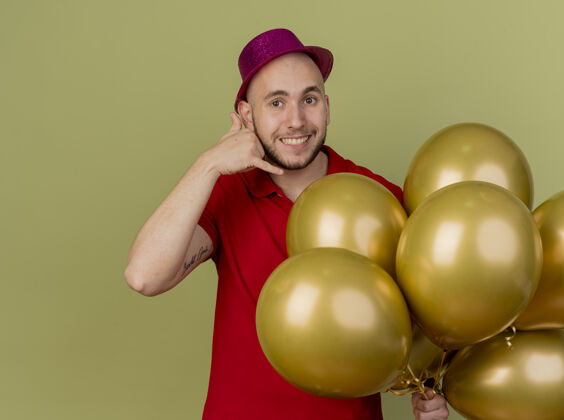 气球微笑的年轻英俊的斯拉夫党人戴着党的帽子拿着气球看着相机做呼叫手势橄榄绿的背景与复制空间隔离帽子微笑抱着