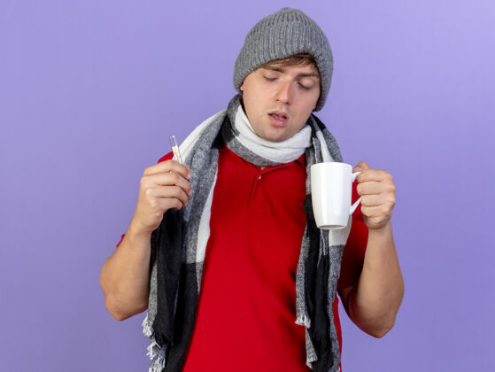 体温计虚弱的年轻英俊的金发病男子戴着冬天的帽子和围巾拿着温度计和杯子隔离在紫色的背景上金发紫色年轻人