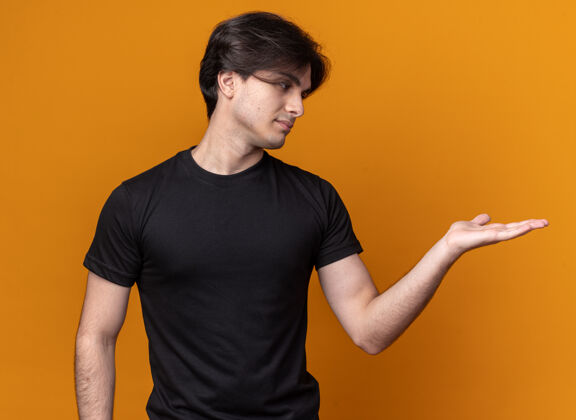 抱着穿着黑色t恤的年轻帅哥假装拿着一个孤立的东西放在橙色的墙上 留着复印空间年轻不愉快假装