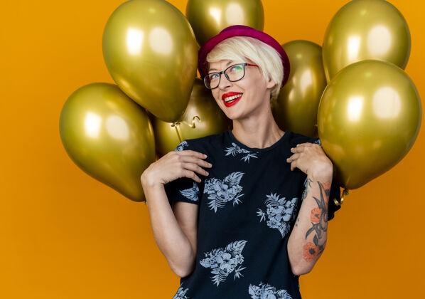 快乐快乐的年轻金发派对女孩戴着派对帽和眼镜站在气球前看着侧面触摸肩膀孤立在橙色背景帽子站气球