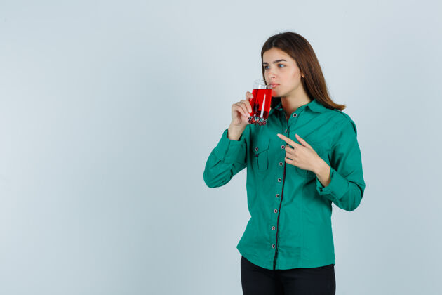 人年轻女孩喝着一杯红色的液体 穿着绿色上衣 黑色裤子 用食指指着它 目光集中前视图饮酒房子室内