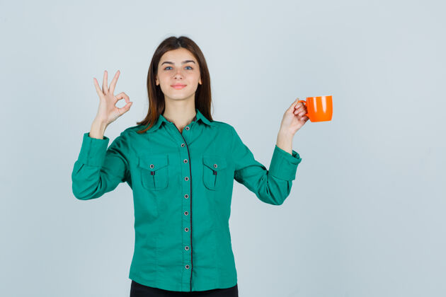 护理年轻的女士拿着一杯橙黄色的茶 穿着ok标志的衬衫 看起来很自信正面图模特而举行