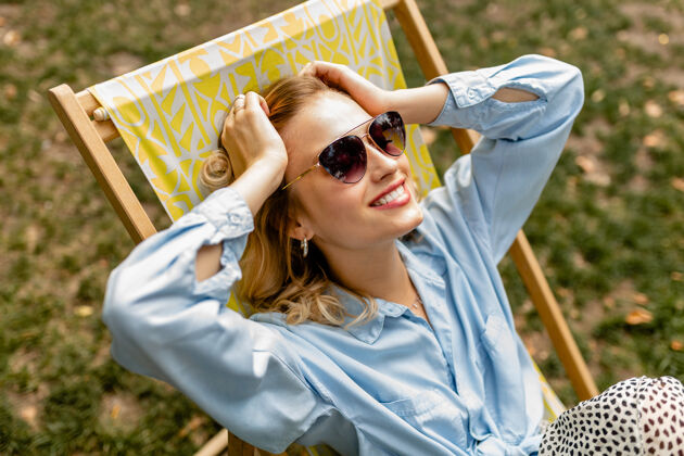明亮迷人的金发微笑的女人坐在躺椅上穿着夏天的衣服夏天放松太阳镜
