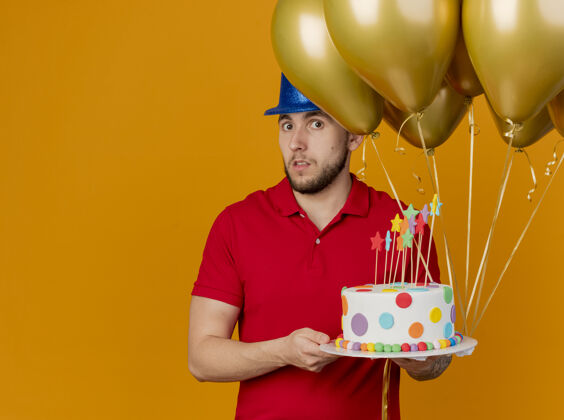 印象令人印象深刻的年轻英俊的斯拉夫党的家伙戴着党的帽子拿着气球和生日蛋糕看着相机孤立的橙色背景与复制空间帽子蛋糕拿着