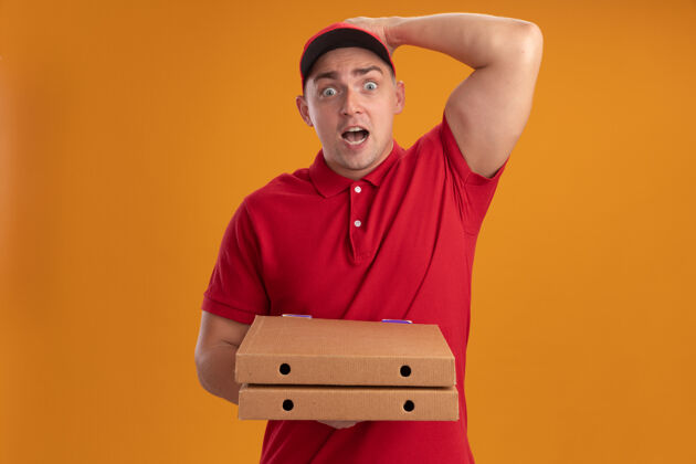 披萨被吓坏了的年轻送货员穿着制服 戴着帽子 拿着披萨盒 手放在隔离在橙色墙上的头上年轻人盒子穿