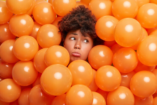 女人沮丧悲伤的非洲头发女人伸出头穿过充气气球悲伤地看着远方不想变老被橙色氦气球包围在生日庆典上孤独沮丧颜色种族