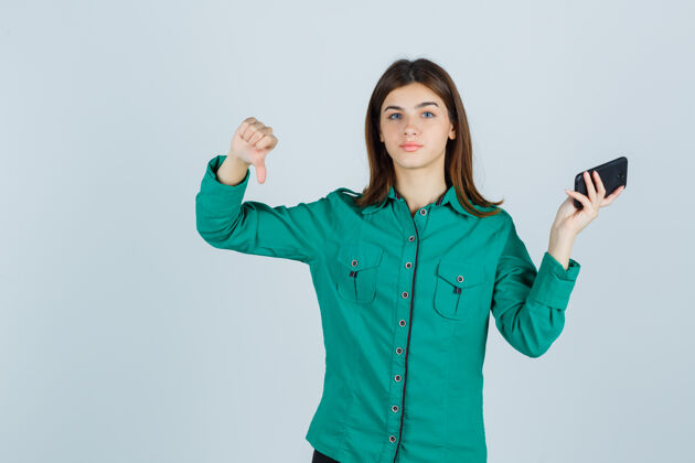 孤独拿着手机的年轻女士 穿着绿色衬衫 大拇指朝下 看起来很不高兴 正前方视图商业不高兴思考