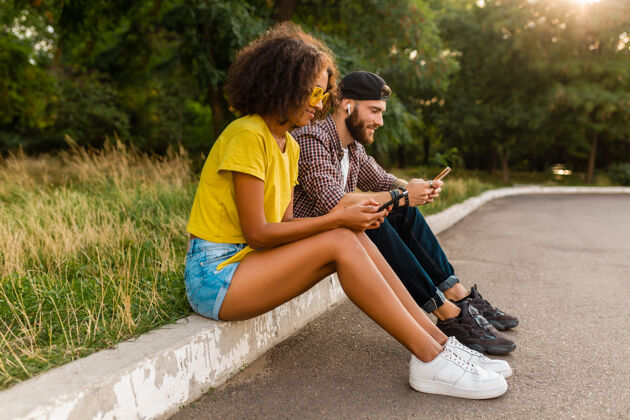 女人快乐的年轻微笑的朋友坐在公园里用智能手机 男人和女人一起玩人酷黑色