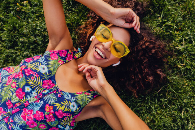 年轻年轻时尚的黑人女子戴着无线耳机听音乐在公园里玩得很开心 夏日时尚风格 五颜六色的时髦装扮 躺在草地上 俯瞰风景混血儿眼镜女性